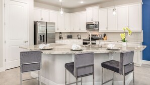 A modern kitchen with white cabinets, stainless steel appliances, a marble island with bar stools, and pendant lighting. - Sophisticated Villa with Movie Room at Encore (Kissimmee)