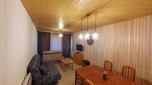 Uma sala de estar aconchegante com teto de madeira, um sofá, uma mesa de jantar de madeira com cadeiras, uma TV e um relógio na parede.