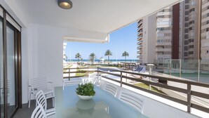Un balcon avec une table en verre, des chaises blanches et une plante en pot. Il donne sur une plage bordée de palmiers, avec un bâtiment visible au loin.