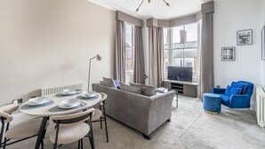 Ein modernes Wohnzimmer mit einem grauen Sofa, einem Esstisch, einem blauen Sessel und einem großen Fenster mit Vorhängen.
