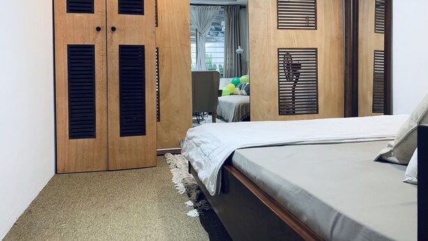 A bedroom with a bed, wooden wardrobe with louvered doors, a small table with a chair, and a view of the outside through a window. - Central Stylish Home (Accra)