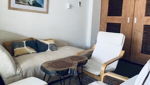 A room with a bed, a white armchair, a small wooden table, and a wardrobe with louvered doors. - Central Stylish Home (Accra)