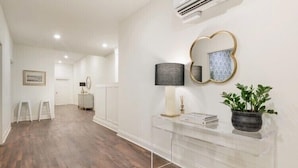 A hallway with a wooden floor, a clear glass console table, a wall-mounted mirror, and a potted plant. - Large, Charming historic flat, Downtown/Bourbon trail/Yum Center (Louisville)