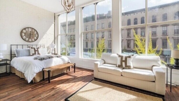 A bedroom with a large window, a bed, a bench, a sofa, and a wall clock. - Large, Charming historic flat, Downtown/Bourbon trail/Yum Center (Louisville)