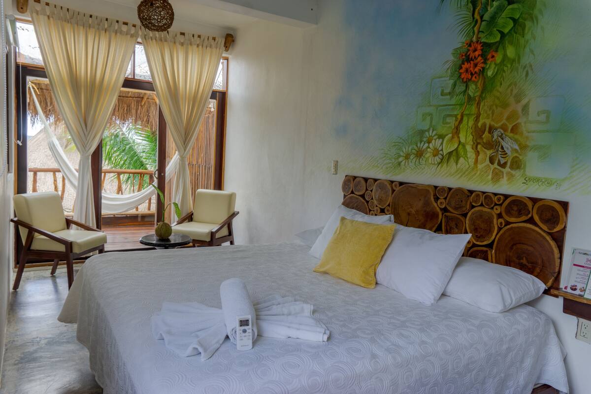 A hotel room with a large bed, a wooden headboard, and a mural of a tropical scene. There are two chairs and a small table with a plant.