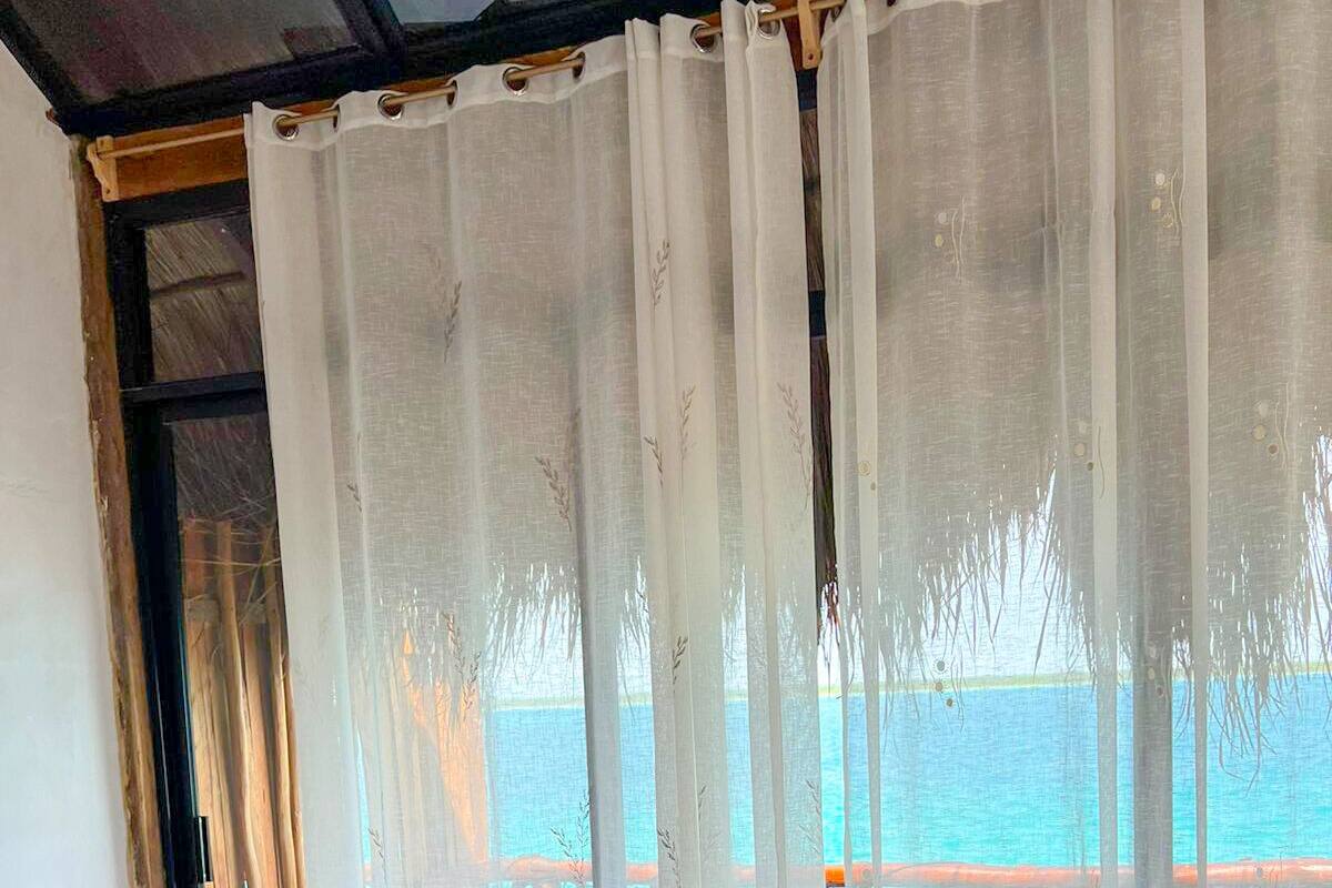 A bedroom with a large bed, white bedding, and a view of the sea through sheer curtains.