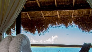 Eine Strandlounge mit einem Reetdach, einem weißen Handtuch, zwei Liegestühlen und einem kleinen Tisch mit Getränken.