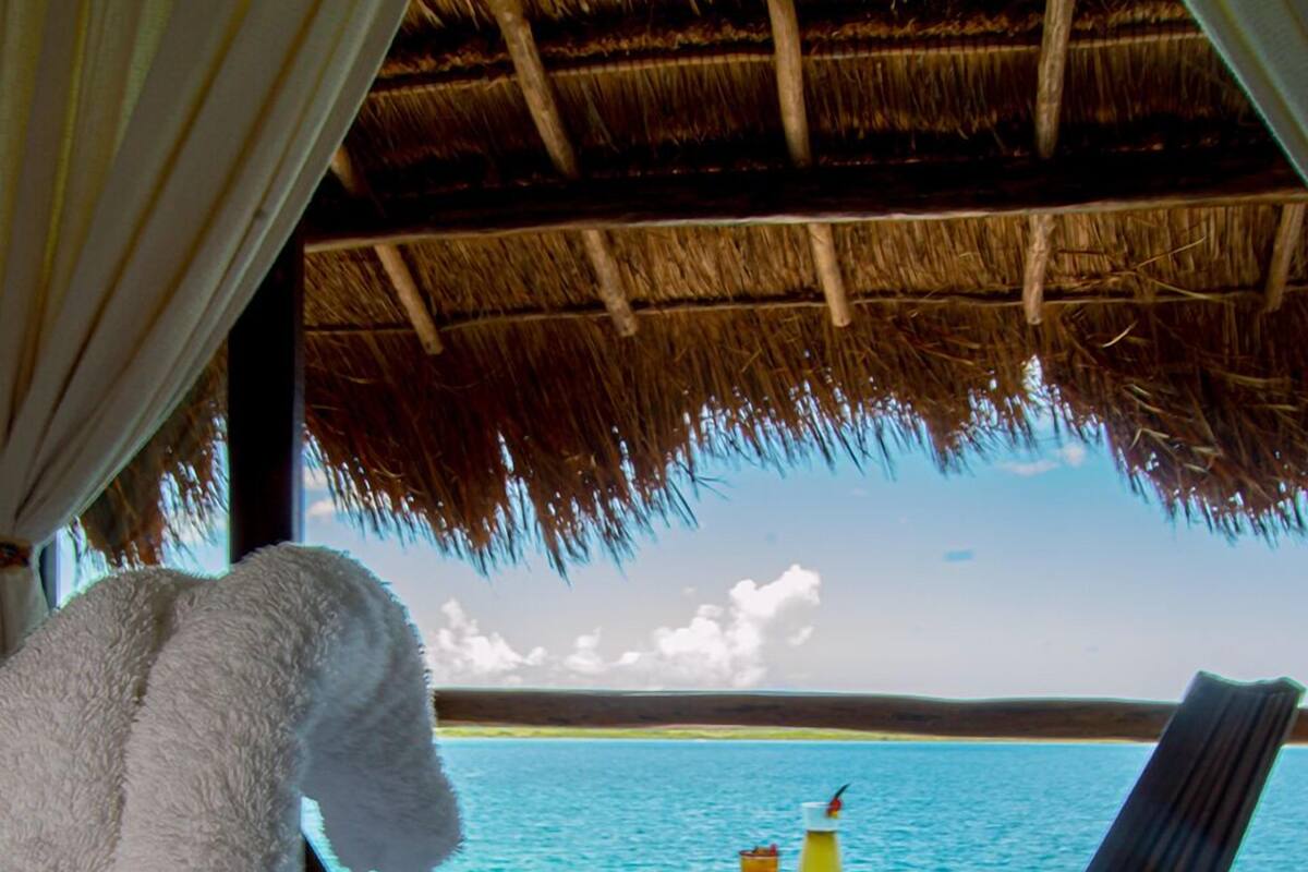 A beachside lounge with a thatched roof, a white towel, two lounge chairs, and a small table with drinks.