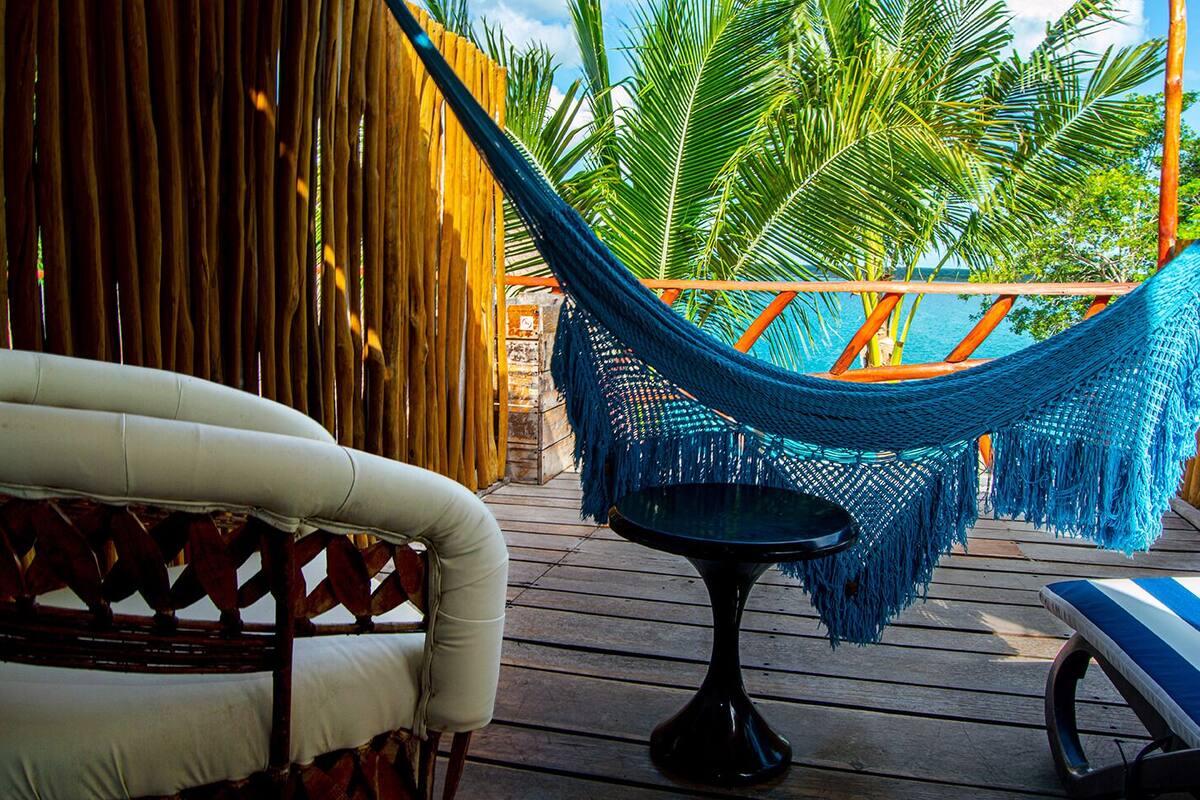 A hammock chair and a small table on a wooden deck with a view of palm trees and a pool.