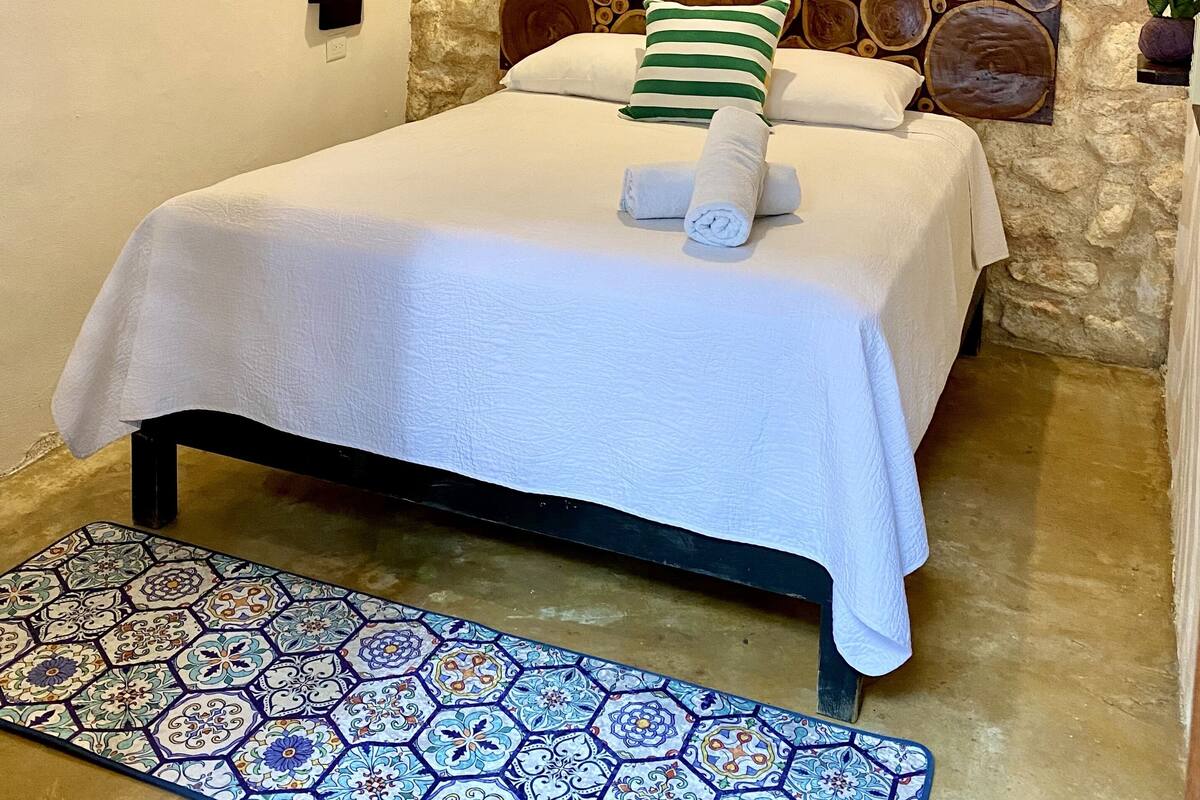 A neatly made bed with white linens, a stone wall behind it, and a small potted plant on the left.