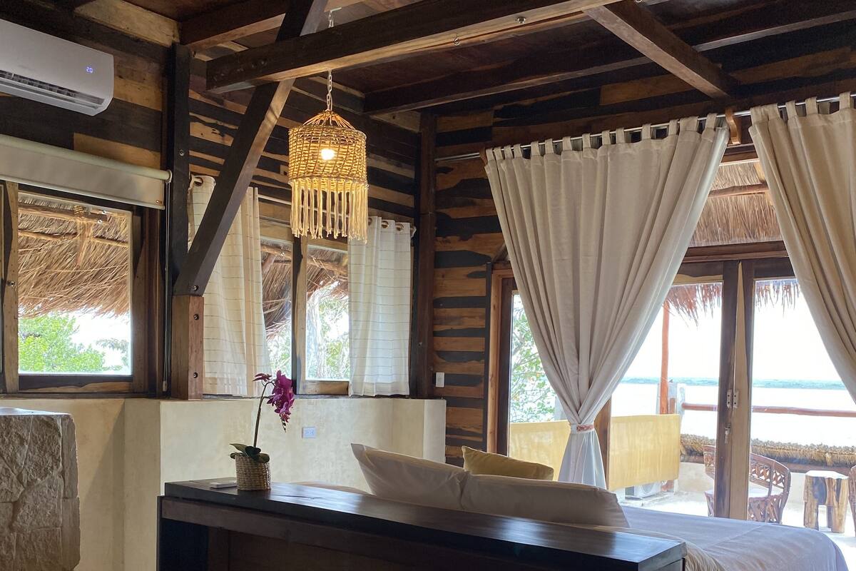 A room with a wooden ceiling, a bed with white bedding, a bedside table with a lamp, and a window with curtains.