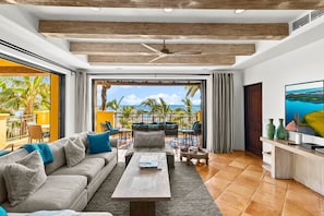 A spacious living room with a large sofa, a coffee table, and a TV on a wooden stand. The room has a wooden ceiling and a view of the beach through large windows. - Newly Renovated Condo with Ocean View & Luxury Amenities in Beachfront Resort. Walk to Downtown (Cabo San Lucas)
