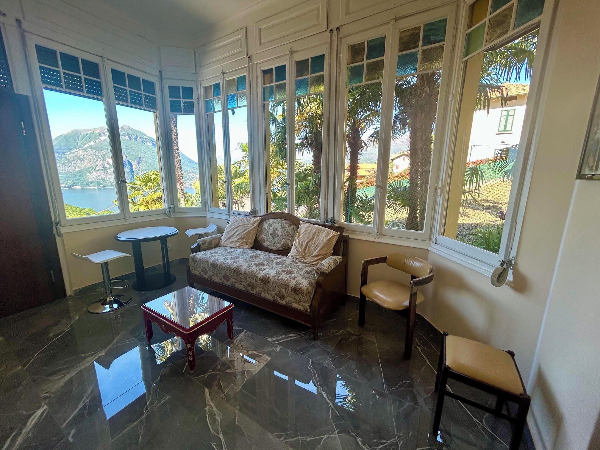 Ein Zimmer mit einer Couch, Sesseln, einem kleinen Tisch und großen Fenstern mit Blick auf Palmen und ein fernes Gebirge.
