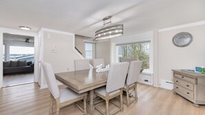 Uma sala de jantar com mesa de madeira e quatro cadeiras, um lustre, um relógio na parede e vista para uma ponte através de uma porta aberta.