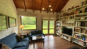 Un accogliente soggiorno con un divano blu, un tavolino da caffè con pedine per gli scacchi, una libreria piena di libri, una televisione e ampie finestre con vista su un paesaggio verde.