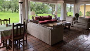 Un accogliente soggiorno con divano, poltrone e tavolo da pranzo. Grandi finestre offrono vista su un rigoglioso giardino.