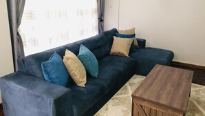 Ein dunkelblaues Sofa mit mehreren Kissen in Blau- und Beigetönen, platziert vor einem Fenster mit einem gemusterten Vorhang. Ein Couchtisch steht davor.