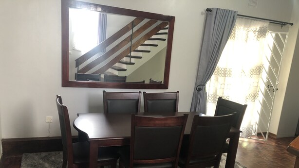 Ein Essbereich mit Holztisch und Stühlen, einem großen Spiegel und einer durch ein Fenster sichtbaren Treppe.