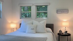 A bedroom with a bed, two windows, a bedside table with a lamp, a wall clock, and a decorative item. - Couples Romantic Country Bungalow, Private, Stock Tank Pool, Birds, Wildlife (Vandalia)