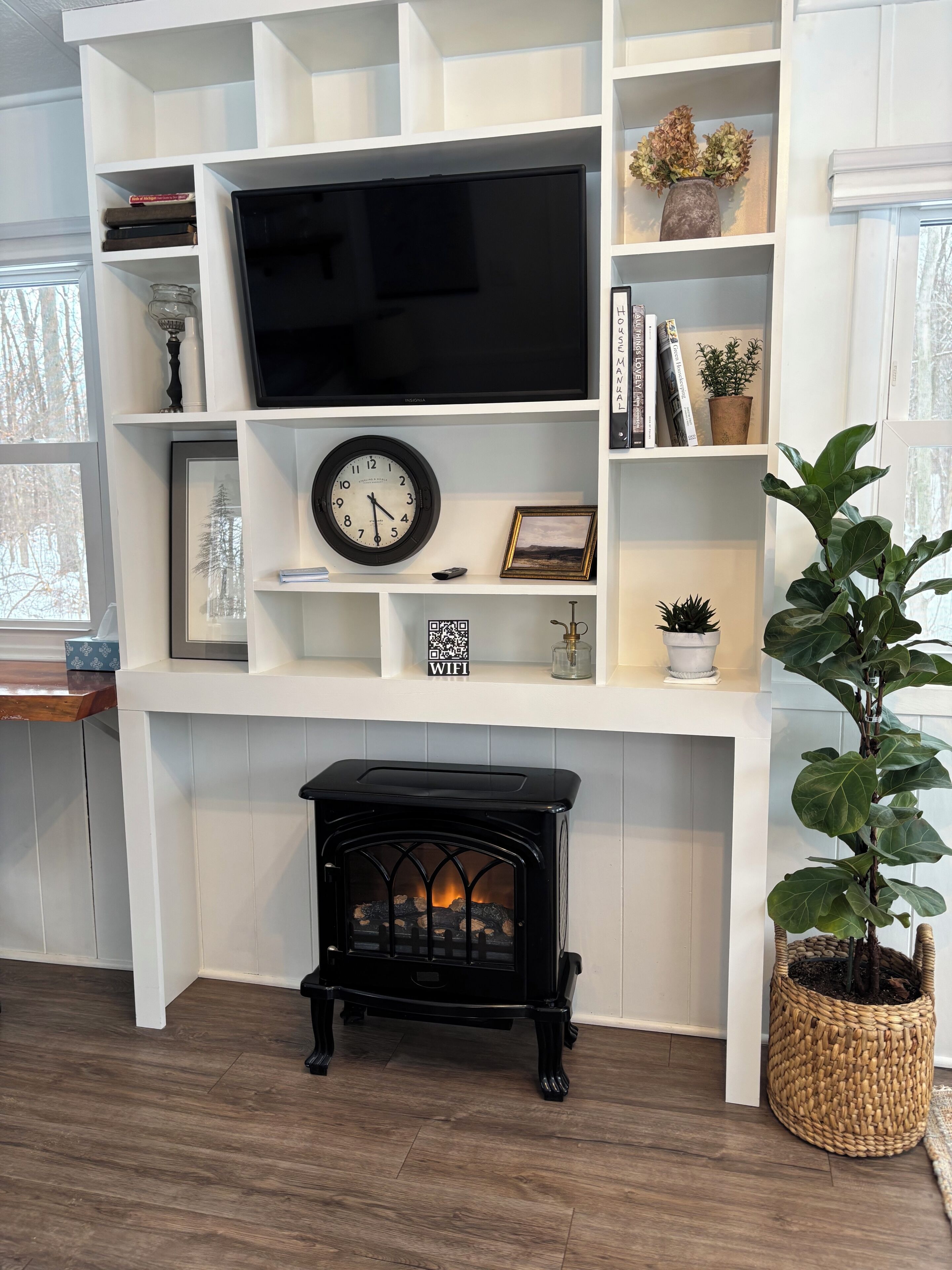 Una sala de estar con chimenea, televisor, un reloj y estantes con libros y objetos decorativos.