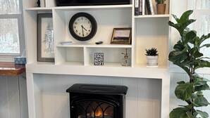 A living room with a fireplace, a television, a clock, and shelves with books and decorative items. - Couples Romantic Country Bungalow, Private, Stock Tank Pool, Birds, Wildlife (Vandalia)