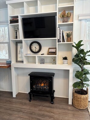 Una sala de estar con chimenea, televisor, un reloj y estantes con libros y objetos decorativos.