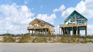 Dos casas frente a la playa con estructuras elevadas y balcones, rodeadas de arena y vegetación.