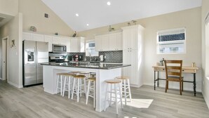 Una cocina moderna con gabinetes blancos, una isla central con taburetes, electrodomésticos de acero inoxidable y un ventanal con persianas.