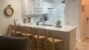 A compact kitchen with white cabinets, a microwave, a refrigerator, and a sink. There are four wooden chairs around a central island. - Orange Beach Oasis  (Orange Beach)