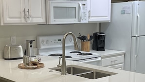 A kitchen with white cabinets, a stainless steel sink, a stove, and a refrigerator. There are three woven stools at the island. - Orange Beach Oasis  (Orange Beach)
