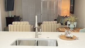 A kitchen with a white countertop, stainless steel sink, and white cabinetry. There is a ceiling fan, a television mounted on the wall, and a window with blinds. - Orange Beach Oasis  (Orange Beach)