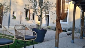 An outdoor seating area with wicker chairs and a table, a potted plant, and a building with windows and a door in the background.