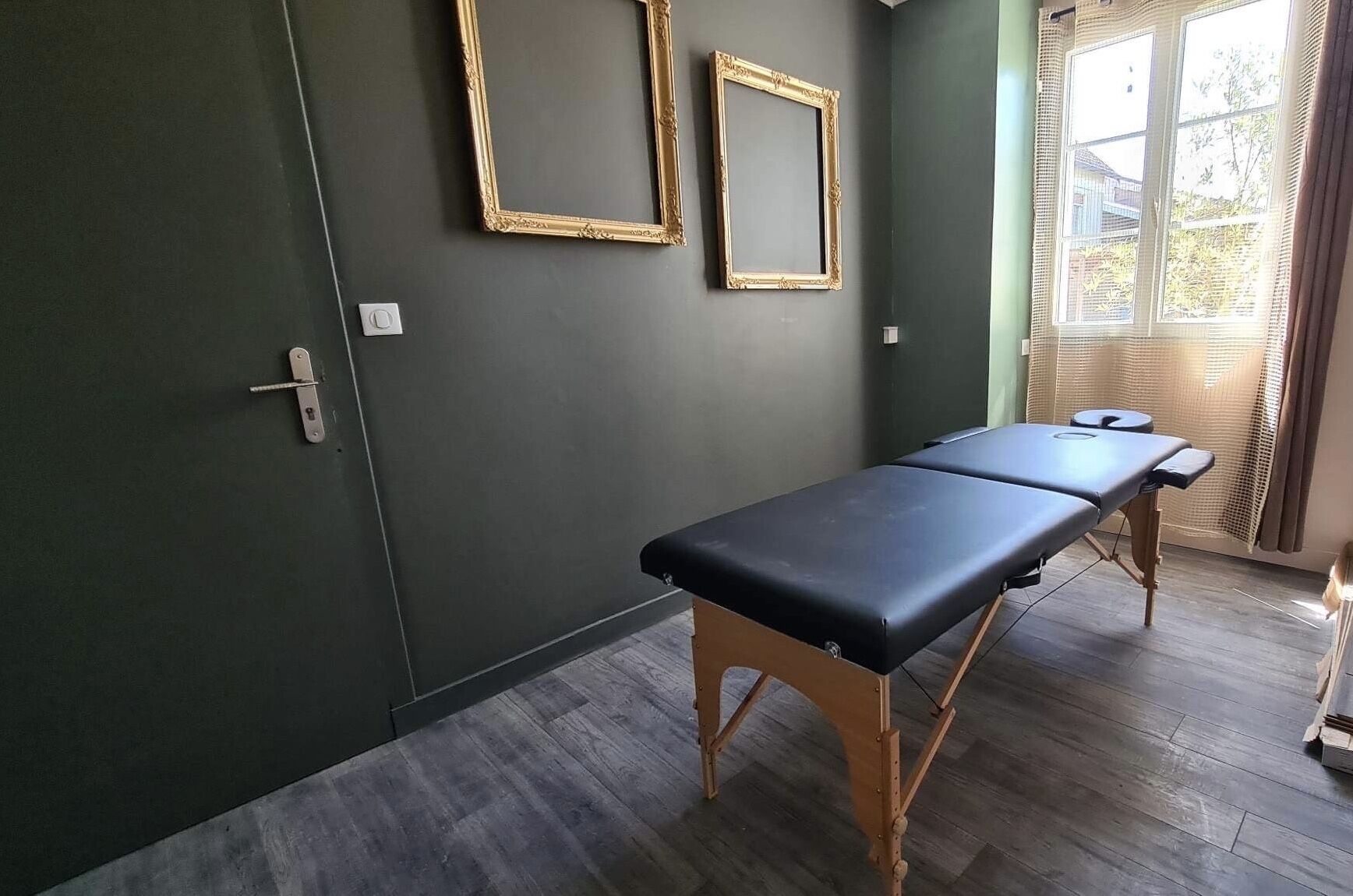 Une pièce avec un mur vert foncé, deux miroirs encadrés de doré, une table d’examen en bois et une fenêtre avec des stores.