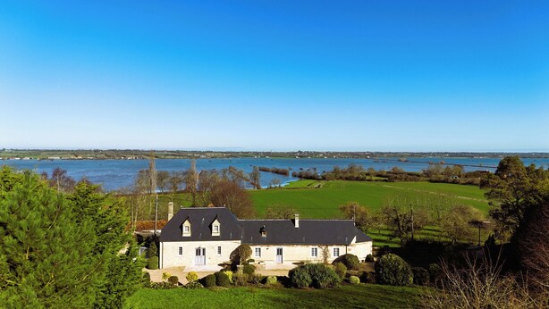 A large stone house with a dark roof, surrounded by lush green fields and a body of water. - Le domaine des roseraies, Omaha Beach, Maupassant gîte (Monfréville)