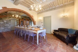 A dining area with a long table, chairs, and a chandelier. There is a sofa and a door in the background.