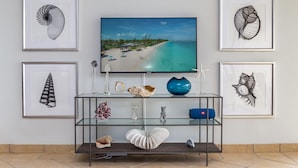 Un salon avec un téléviseur fixé au mur, entouré d’œuvres d’art encadrées, une table basse en verre agrémentée d’objets décoratifs et un sol carrelé.