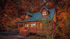 A wooden cabin with a porch and a second-story balcony, surrounded by autumn foliage. - Wooded Smokies Hideaway • Arcade, Pool Table & Hot Tub (Sevierville)