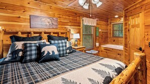 A cozy wooden cabin bedroom with a bed, a nightstand, a lamp, a window with blinds, and a ceiling fan. - Wooded Smokies Hideaway • Arcade, Pool Table & Hot Tub (Sevierville)