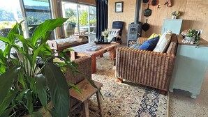Un salon chaleureux avec une grande plante, des meubles en osier, un tapis à motifs et une vue sur l’extérieur.