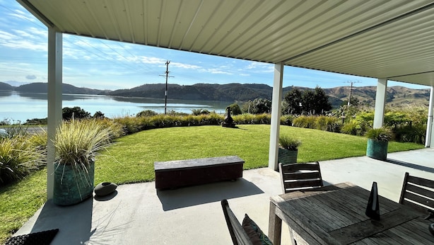 Une terrasse couverte avec vue sur un lac, une pelouse verdoyante et un paysage vallonné en arrière-plan.