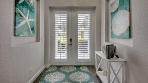 A hallway with a white door, two windows with blinds, a white console table with a statue, and artwork on the walls.