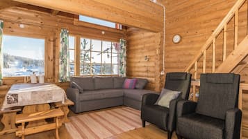 Interior de una cabaña de troncos con sofá, sillón, mesa de madera y vistas a un paisaje nevado.