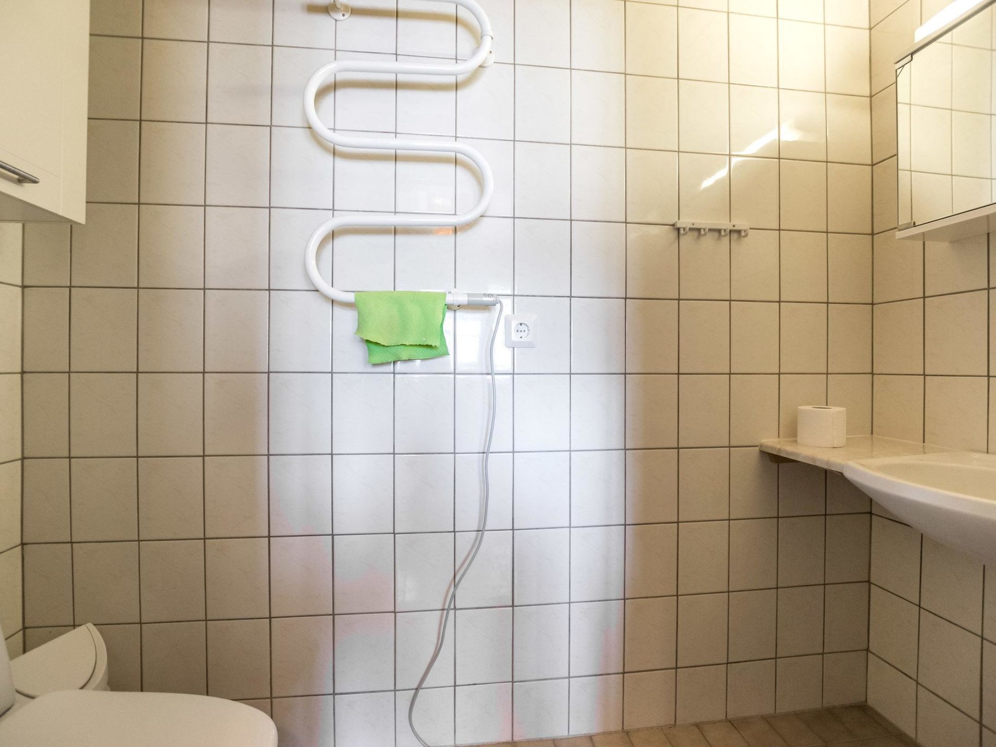 Een badkamer met witte betegelde wanden, een toilet, een douche met een witte slang en een groene handdoek hangend aan een haak.