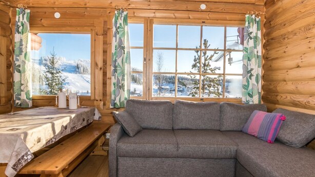 L’intérieur d’une cabane en rondins, avec un canapé, un banc en bois et une fenêtre donnant sur un paysage enneigé.