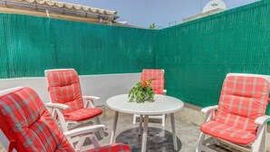 Eine Terrassenfläche mit vier rot-weiß karierten Stühlen um einen weißen Tisch, ein grüner Zaun im Hintergrund und eine Satellitenschüssel auf dem Dach.