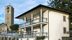 Ein zweigeschossiges Gebäude mit einem Balkon im oberen Stockwerk, einem dahinterliegenden Stein-Turm und blauem Himmel.