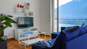 Ein modernes Wohnzimmer mit einem Flachbildfernseher, weißen Regalen, einem Glastisch, einem blauen Sofa und einem großen Fenster mit Blick auf die Berge.