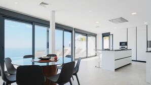 Una cocina moderna con gabinetes blancos y una isla grande, un comedor con mesa y sillas de vidrio, y vistas al océano a través de ventanas amplias.