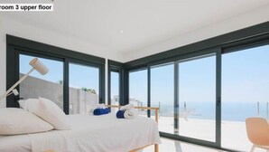 Un dormitorio con cama, mesita de noche, una silla y ventanas amplias con vistas al mar.