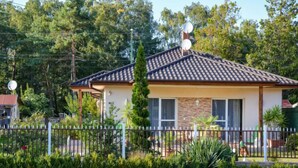 Ein kleines Haus mit Ziegeldach, Satellitenschüsseln und einem umzäunten Garten mit verschiedenen Pflanzen.